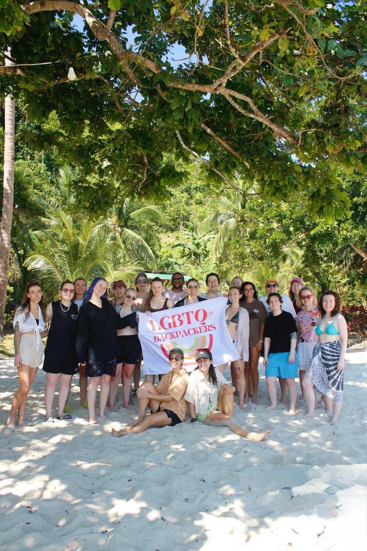 Large backpacker group on a tropical beach hold an LGBTQ+ flag amid palms and bright sunshine.