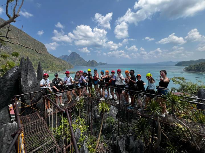 Helmeted travellers stand on a jagged limestone viewpoint overlooking turquoise sea and dramatic karst islands.