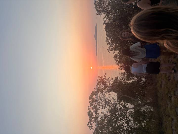 Travellers watch an orange sun set over calm sea from a grassy hilltop.