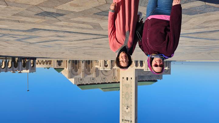 Two women stand before the vast forecourt of Casablanca’s Hassan II Mosque on a clear blue morning.