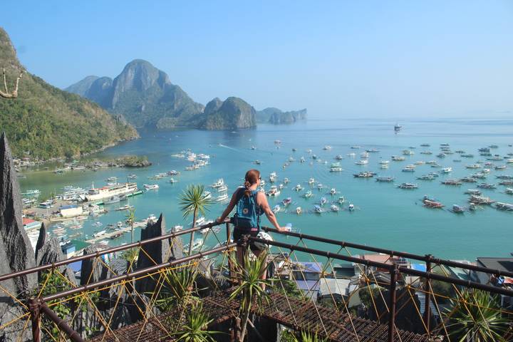 Solo traveller looks out over El Nido harbour packed with boats from a high rocky viewpoint.
