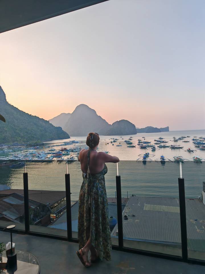Traveller enjoys pastel sunset views of El Nido’s bay and karst formations from a glass balcony.