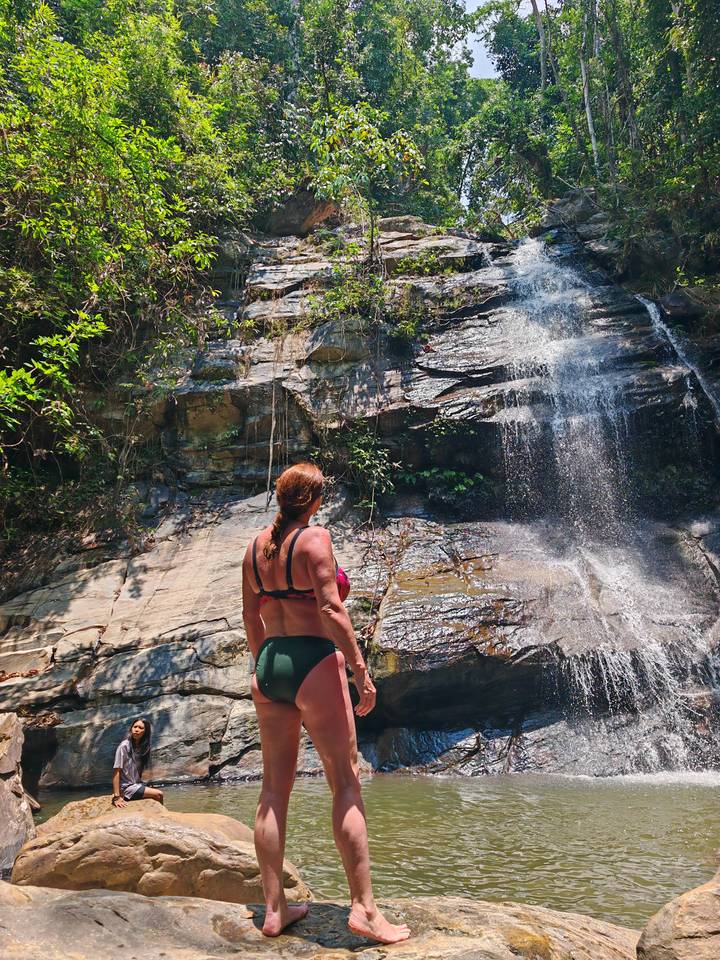 Woman in swimwear stands at the base of a small jungle waterfall admiring the cascading water.