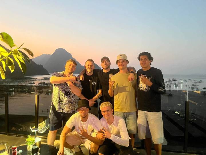 Group of young men pose on a rooftop terrace at dusk with El Nido bay and limestone peaks behind.