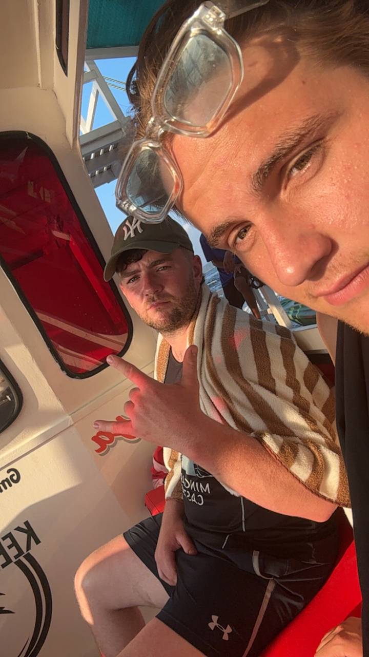 Close-up boat selfie of two young men wrapped in a towel with sea and sunset light in background.