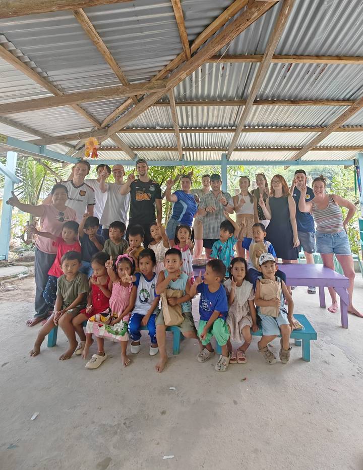 Large group of travellers and village children gather for a joint photo under a corrugated roof.