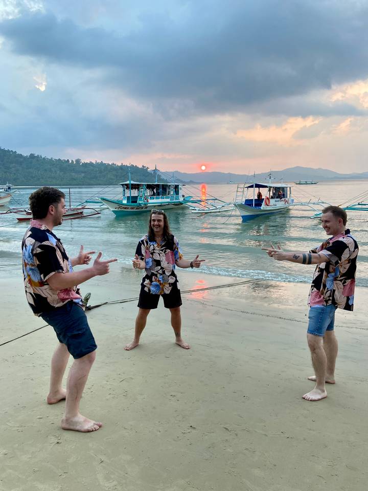 Three men in matching tropical shirts joke around on a beach at sunset with boats behind them.