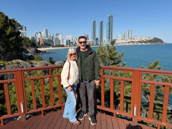 Two travellers smile on a wooden lookout with Busan’s skyscraper-lined coastline behind.