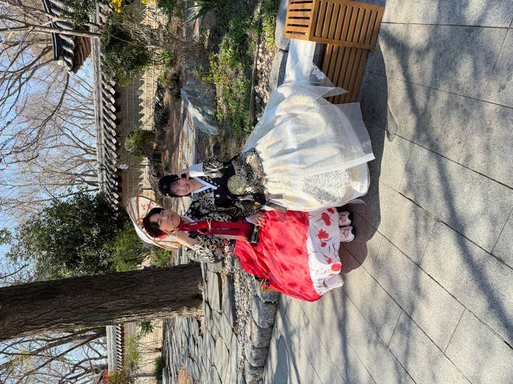 Two women in colourful hanbok pose on a bench beneath bare trees in a hanok village.