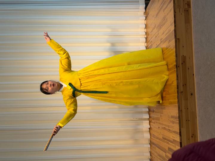 Performer in bright yellow hanbok sings passionately on a wooden stage.