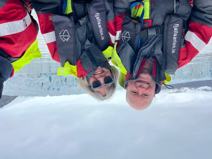 Two travellers in bright flotation suits smile on a glacier lagoon boat with ice wall behind.