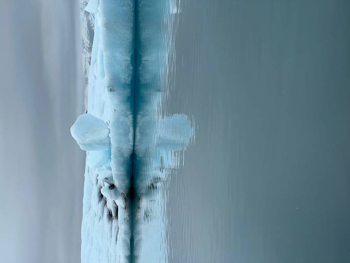 Solitary iceberg reflects in still milky-blue glacial water under grey skies.