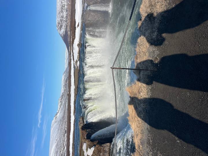Mighty Godafoss waterfall thunders over a horseshoe cliff as onlookers’ shadows fall in foreground.