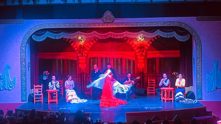 Indoor flamenco performance on a warmly lit stage with a dancer in a red dress and musicians seated behind.