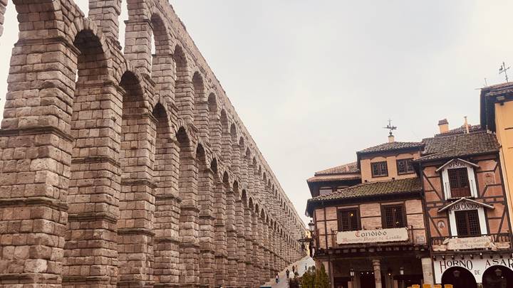 Side perspective of ancient Roman aqueduct arches beside traditional Spanish buildings.