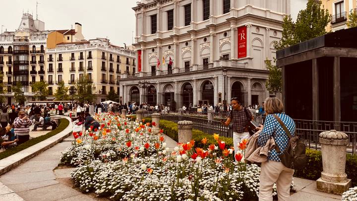 Busy urban plaza with colorful tulip beds, pedestrians, and a grand neoclassical building.