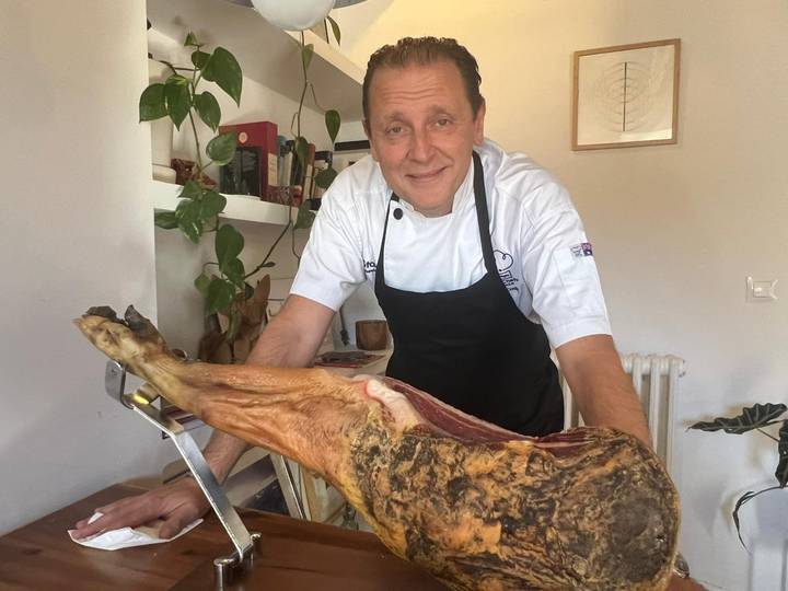 Chef proudly presents a leg of cured ham on a slicing stand inside a cozy kitchen.