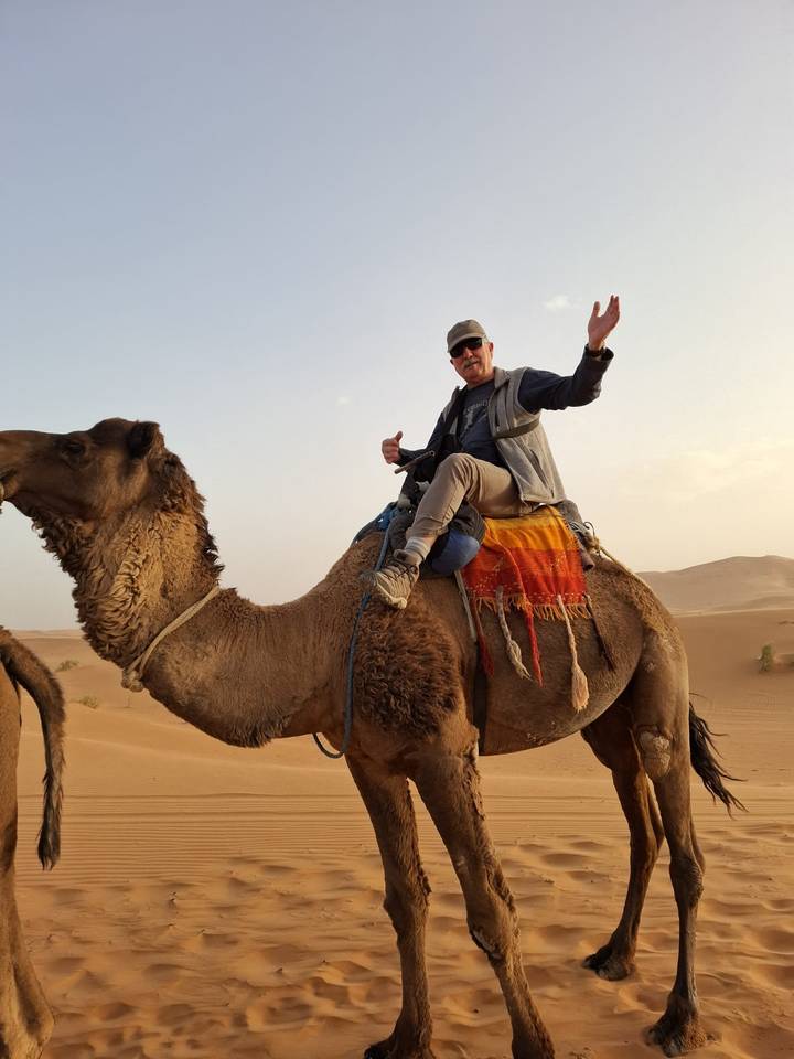 Traveler riding a camel draped with a colorful blanket across rolling desert dunes at dusk.