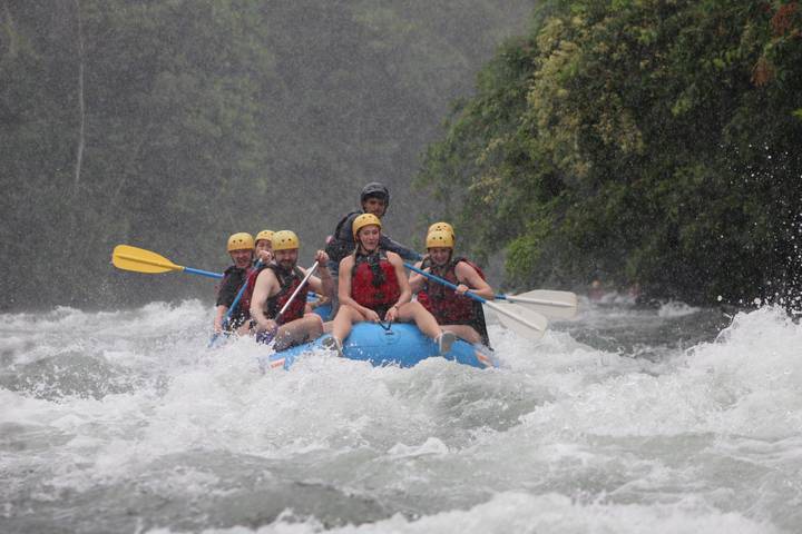Raft with excited paddlers plunging through white-water rapids amid jungle river scenery.