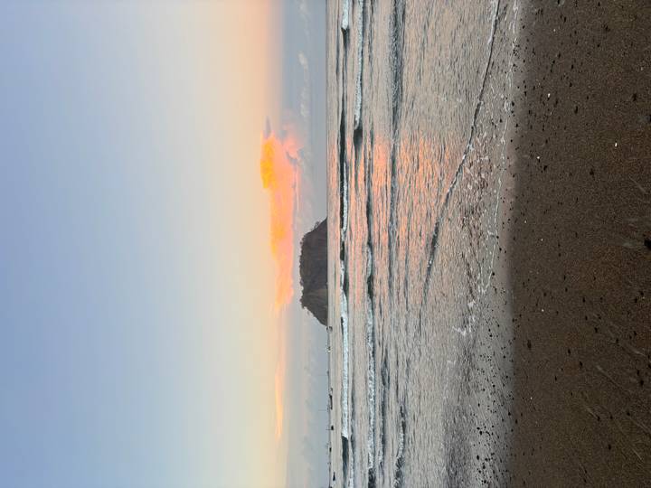 Island silhouette under pastel sunset skies reflected on calm coastal waters from a sandy shoreline.