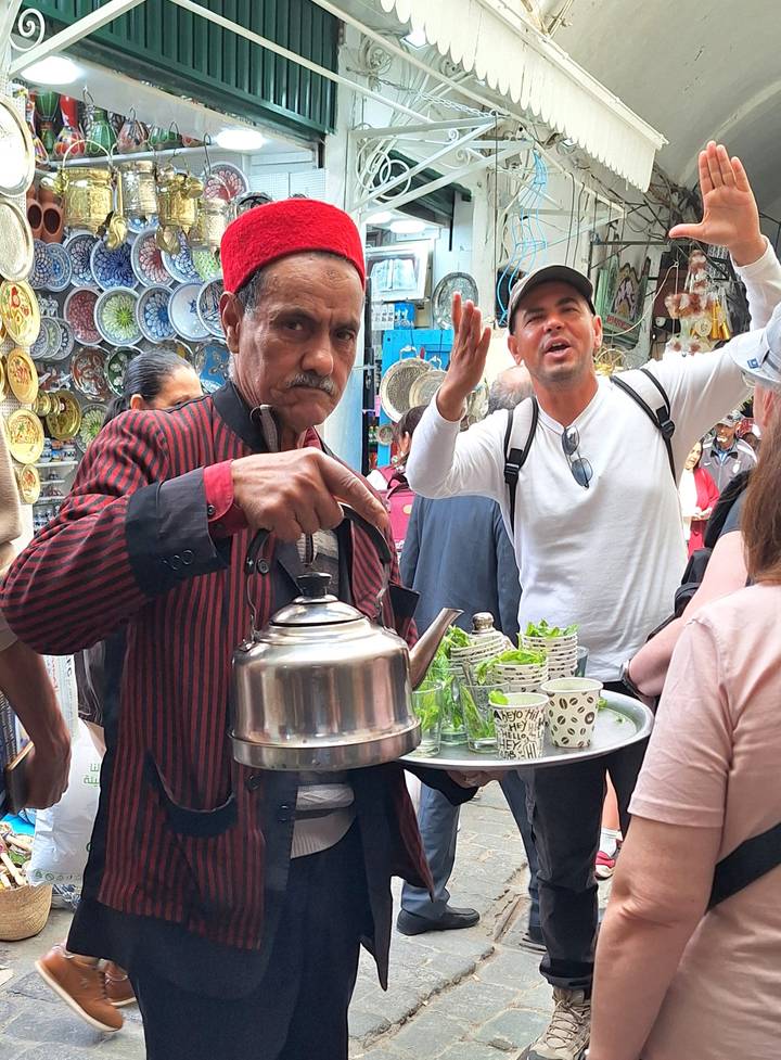 Street vendor presenting a metal teapot to curious shoppers in a bustling Tunis medina market.