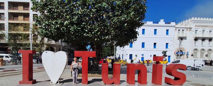 Large red letter sculpture spelling 'I ♥ Tunis' with a tourist posing beside it in a sunny plaza.