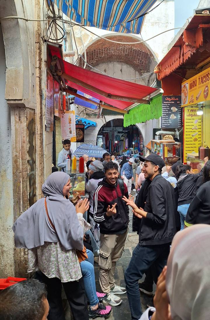 Crowded medina alley with vibrant signs, food stalls and animated locals chatting.
