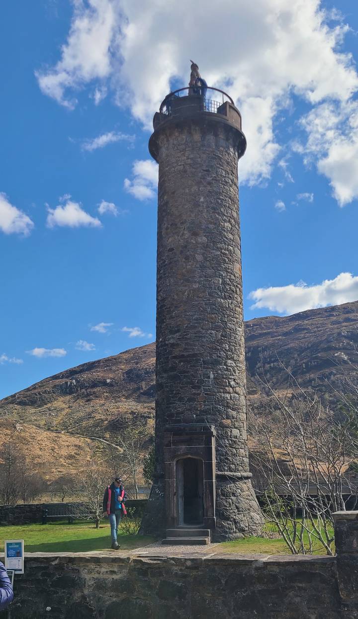 Stone Glenfinnan Monument tower rising beside a loch with rugged hills and blue sky.