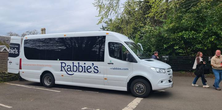 White Rabbie’s tour minibus parked under leafy trees with travellers nearby.