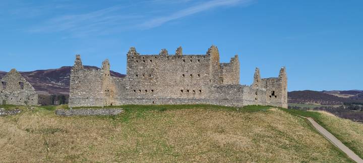 Ruthven Barracks stone ruins stand isolated on a grassy mound with mountains behind.