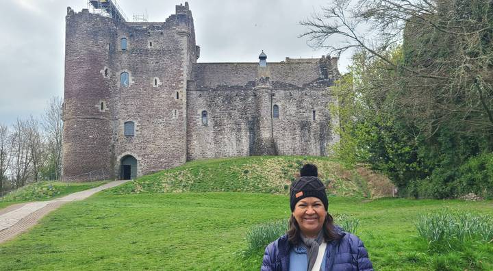 Smiling traveller stands before the imposing grey stone walls of Doune Castle surrounded by greenery.