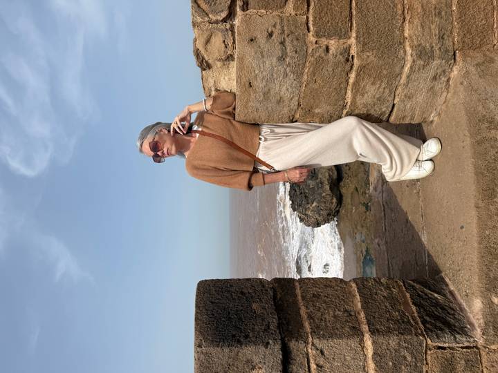 A stylish woman leans against an old seaside stone wall gazing over waves under a clear sky