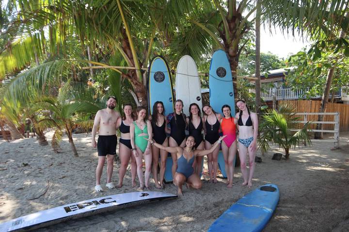 Surf school group posing with longboards under palm trees on tropical sand.