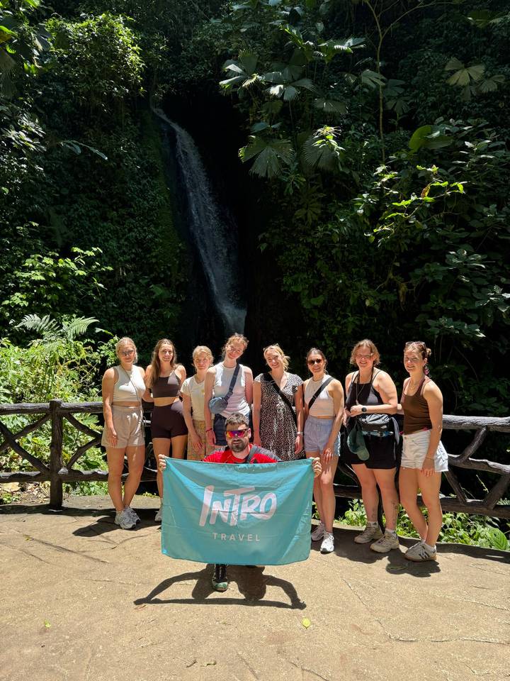 Tour group standing on a wooden bridge in front of a lush jungle waterfall, holding a banner.