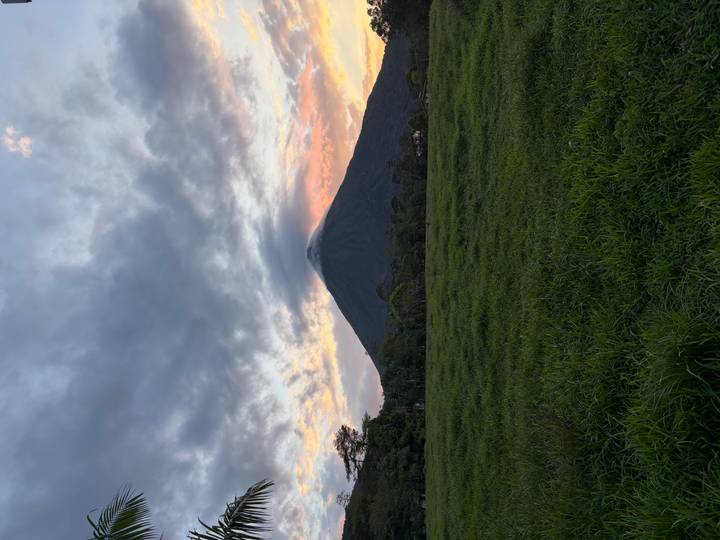 Dramatic view of Arenal Volcano rising above green pastures under colorful dawn clouds.
