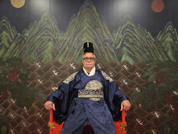 Man dressed in royal Joseon robe seated regally before a painted mountain throne backdrop.