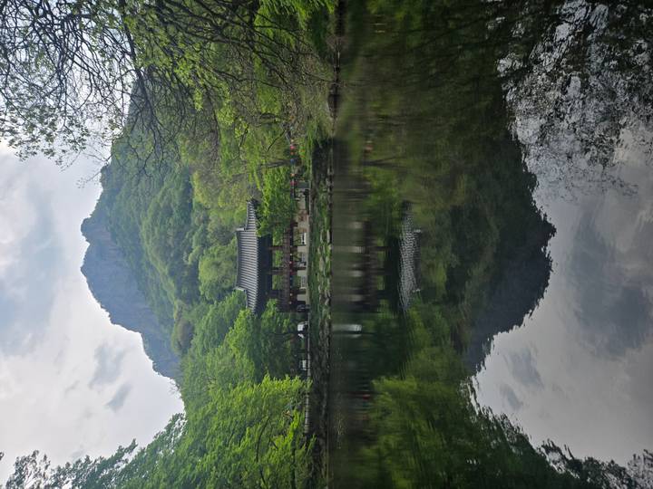 Serene pond reflecting a traditional pavilion beneath a steep forested mountain cliff.