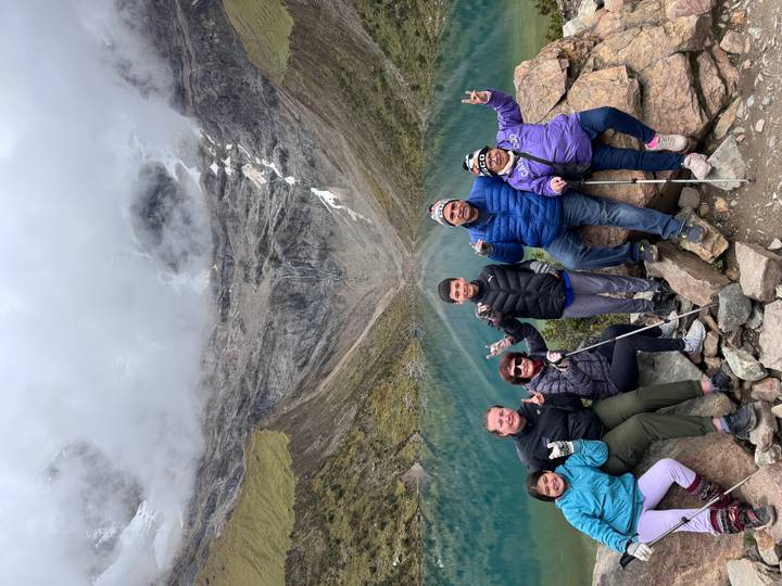 Family of hikers posing triumphantly on rocks above the turquoise Humantay Lake and glacial mountain walls.