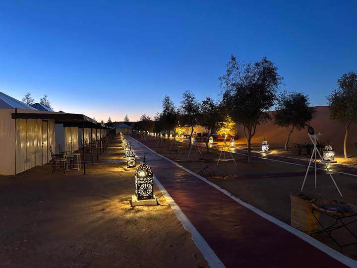 Evening view of a luxury desert camp walkway lined with glowing lanterns between tents and dunes.