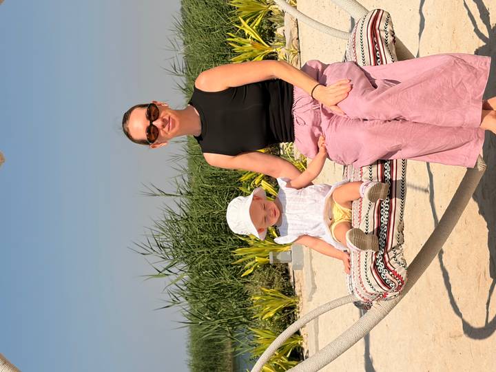 Woman and toddler sitting on a patterned hammock over white sand with tall reeds and blue sky behind.