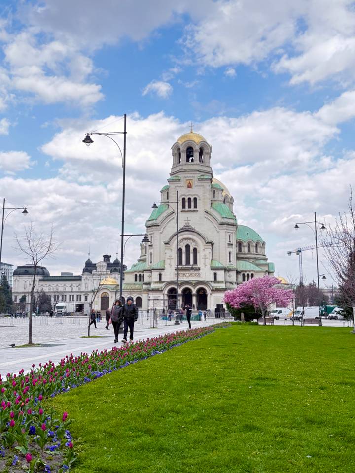 Grand neo-Byzantine cathedral with green domes and arched façade in a city square with pedestrians and blooming trees.