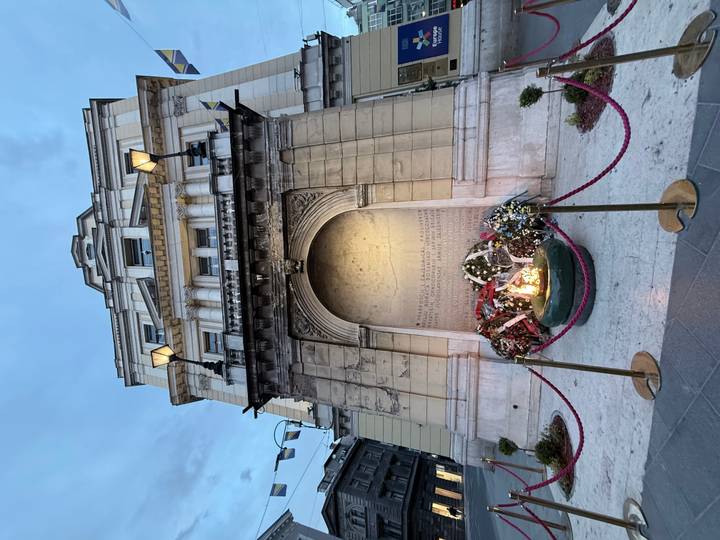 Stone arch memorial with eternal flame and wreaths in front of a historic building at dusk.