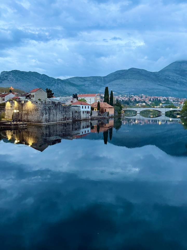 Calm river reflecting stone walls, red-roofed houses and an arched bridge with mountains in the background.