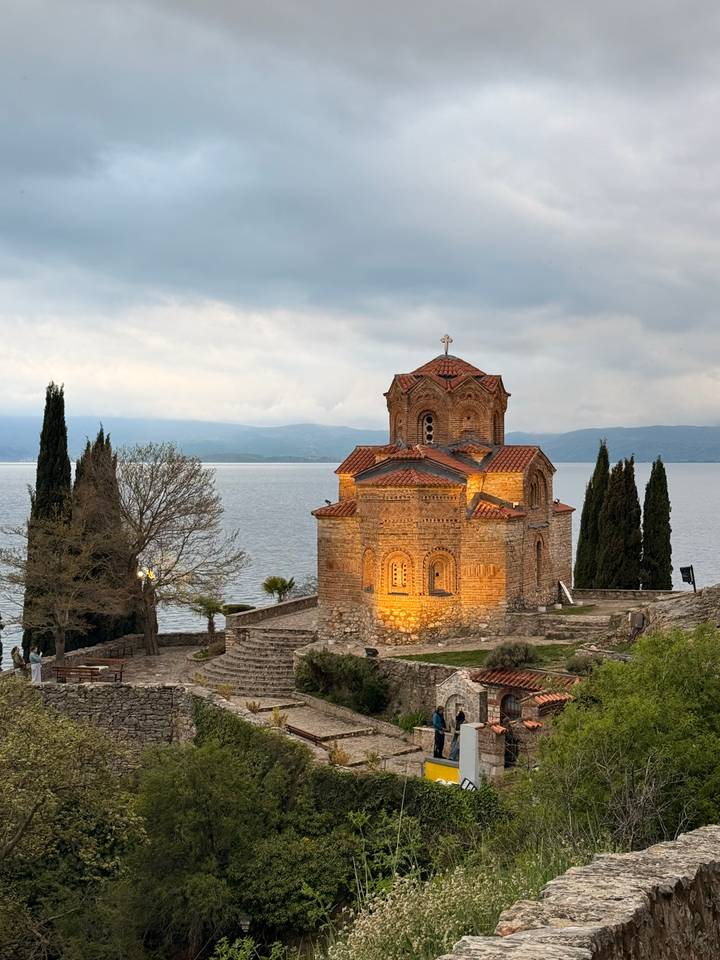 Picturesque Byzantine church perched on a rocky bluff above a vast lake with mountains under moody skies.
