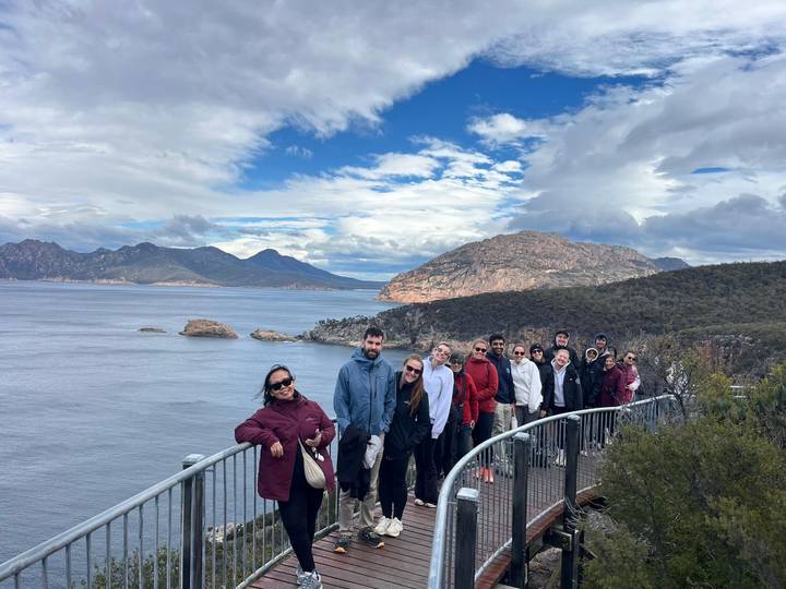 Tour group posing on a coastal lookout railing with sweeping views of a blue bay and rugged mountains.