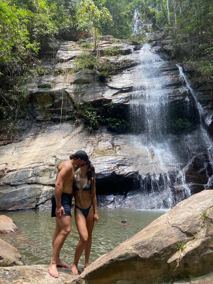 Couple in swimwear shares a kiss beneath a tropical waterfall cascading over smooth rock.