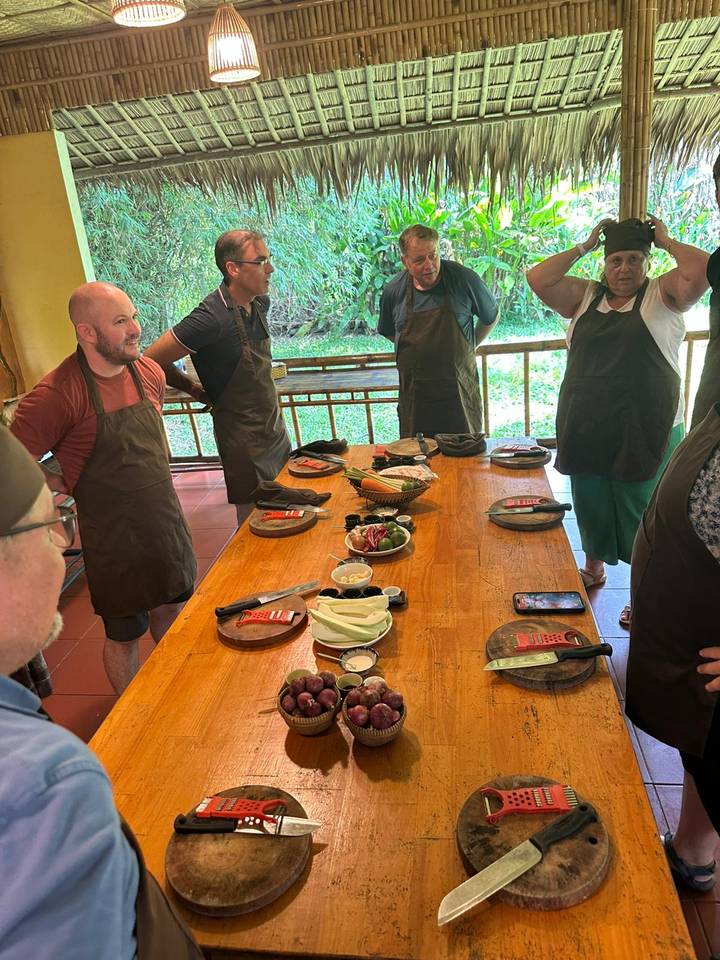Cooking class participants wearing aprons stand around a wooden table set with ingredients and utensils.