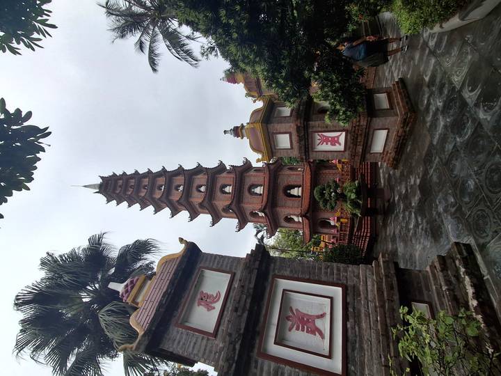Tall red-brick multi-tiered pagoda surrounded by small shrines and greenery on an overcast day.
