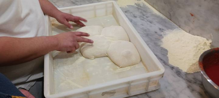 Chef’s hands portioning fresh pizza dough in a plastic tray on a marble counter.