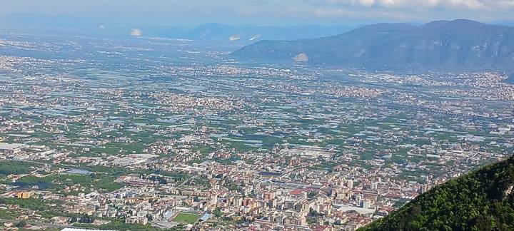 Zoomed aerial view of an Italian town and patchwork farmland with mountains beyond.